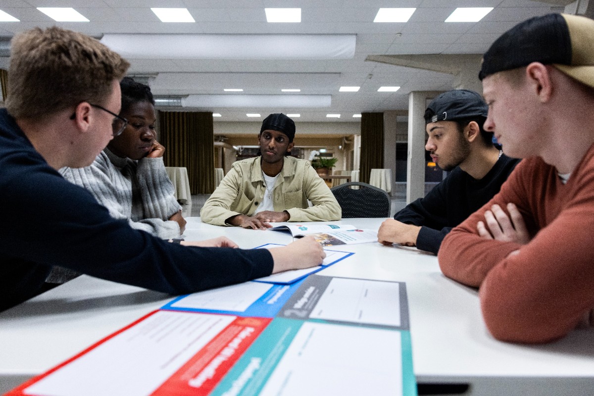 Fem unga personer sitter vid ett bord och för dialog.